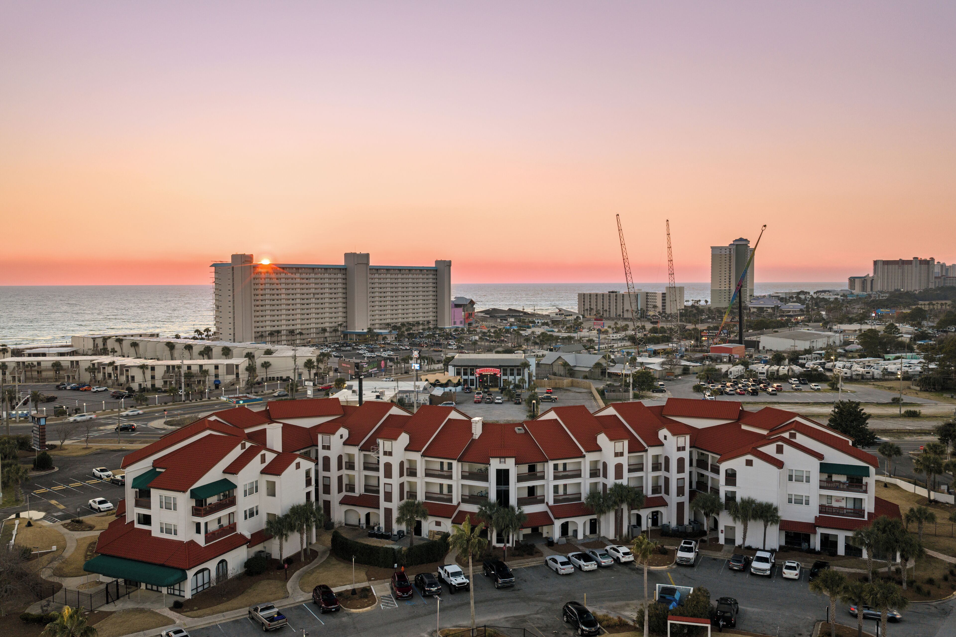 A multi-story hotel with a red roof, located near a beach with a sunset in the background.