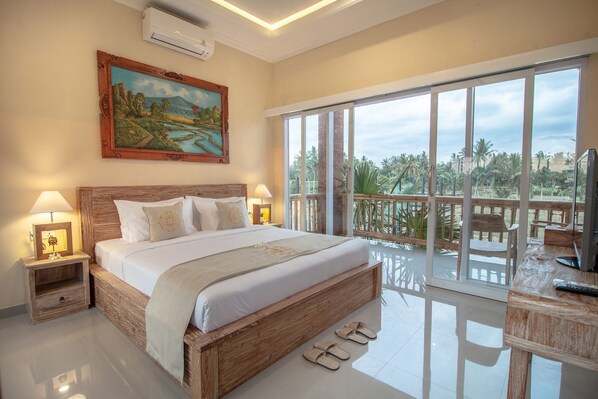 A bedroom with a large bed, two bedside tables with lamps, a painting on the wall, and a balcony with a view of palm trees. - Sujiwa Ubud Retreat (Sukawati)
