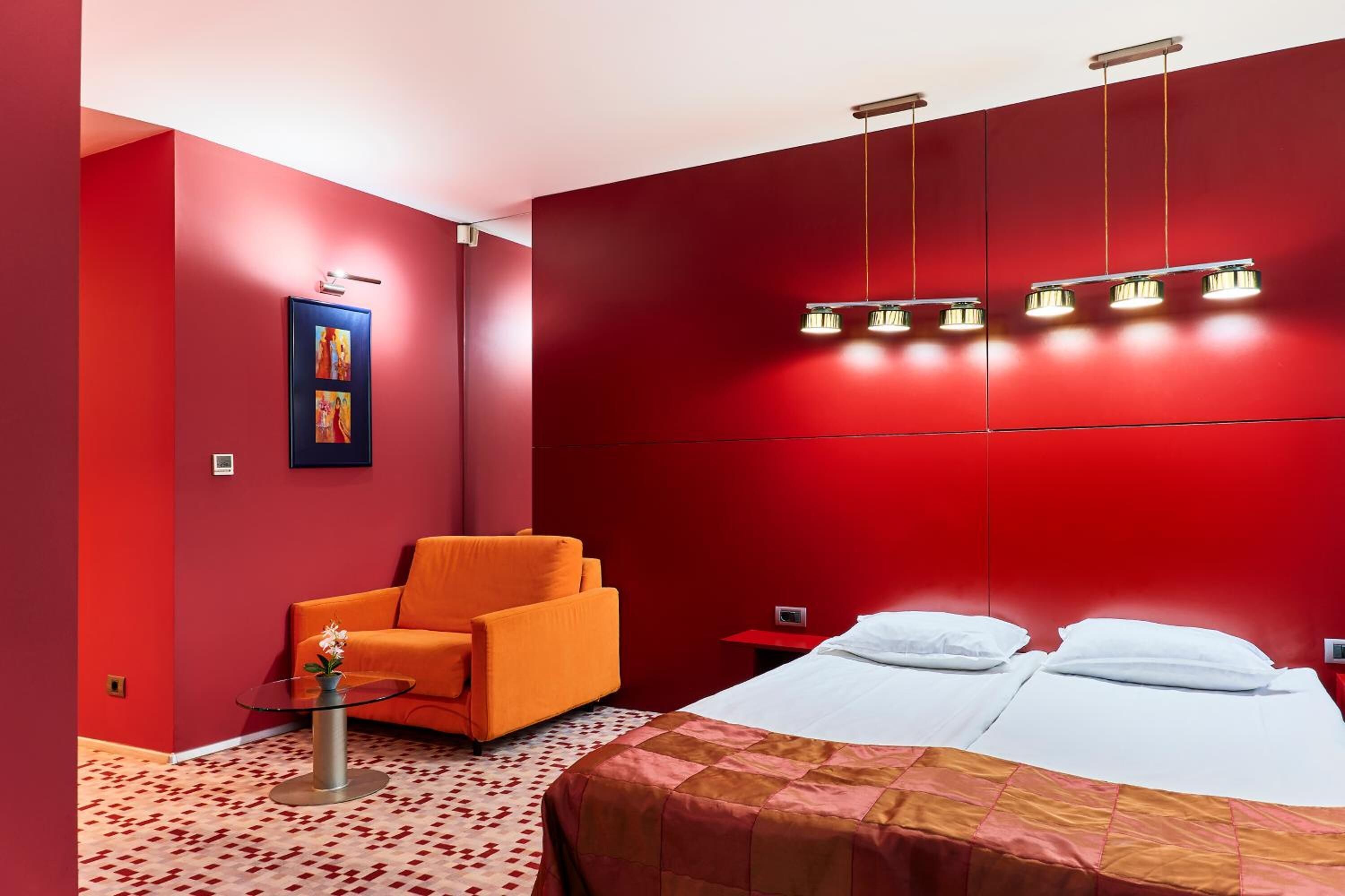 Una habitación de hotel con pared roja, una cama con ropa de cama blanca, un sillón naranja, una mesita redonda con un florero y una lámpara de techo.