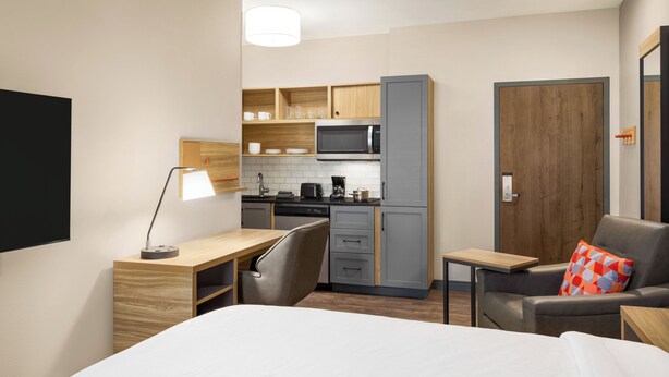 A compact hotel room with a kitchenette, a bed, a desk with a lamp, and a seating area with a couch and a pillow. - Woodspring Suites Pecos (Pecos)
