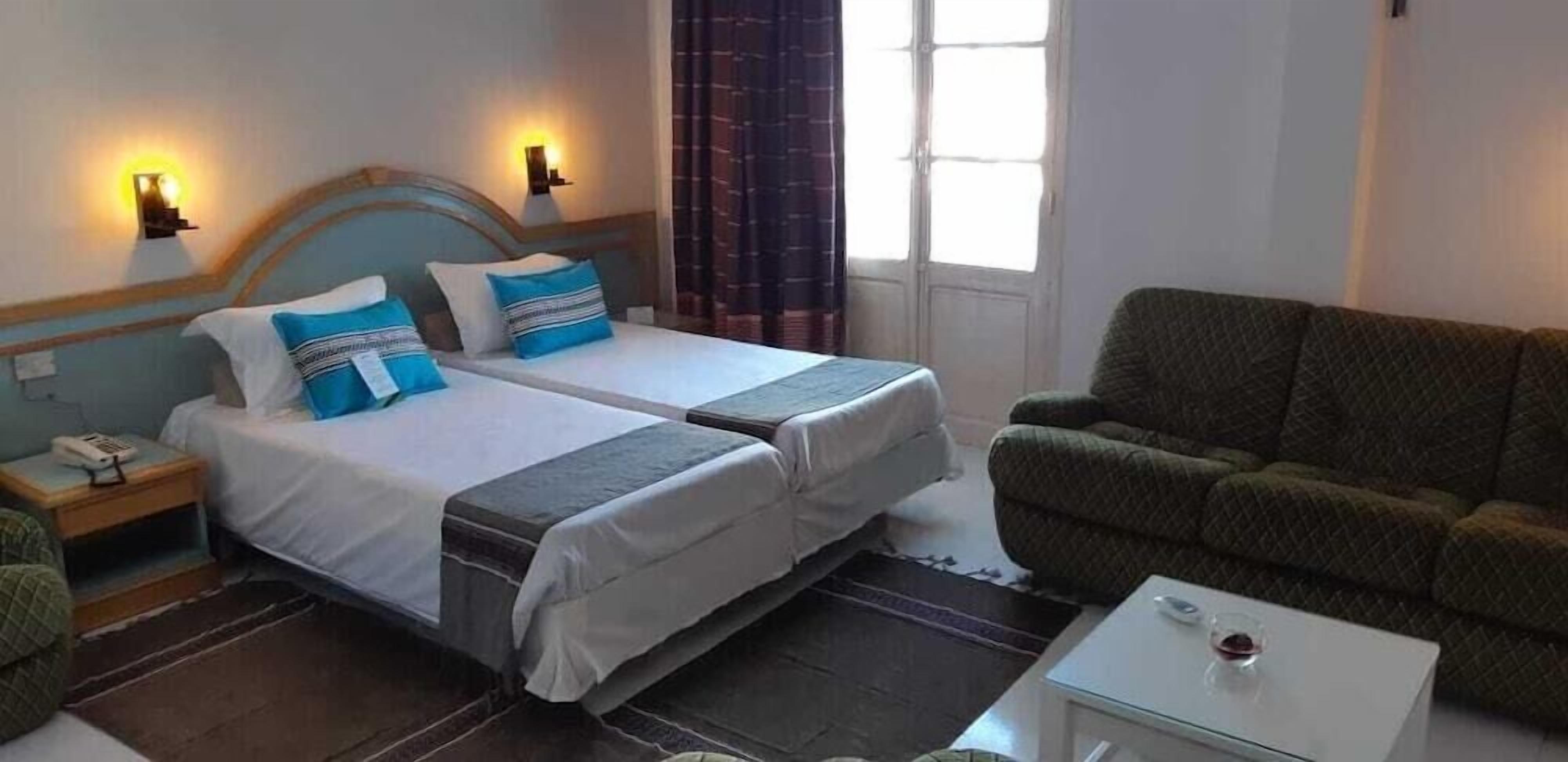 A hotel room with two beds, a green sofa, a small table with a glass, and a window with curtains.
