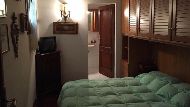 Une chambre avec un lit vert, une armoire en bois, un petit bureau avec une télévision et un luminaire fixé au mur.