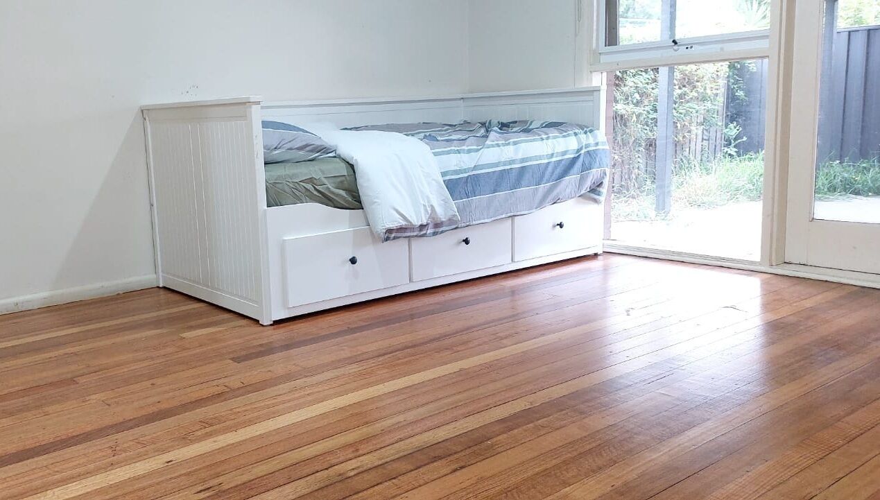 A room with a white bed in the corner, wooden flooring, and a large window providing a view of the outdoors.