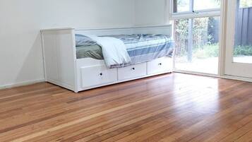 A room with a white bed in the corner, wooden flooring, and a large window providing a view of the outdoors.