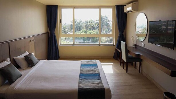A hotel room with a large bed, a desk, a chair, a TV, and a window with a view of palm trees. - Cosmique Clarks Inn Suites Goa (Margao)