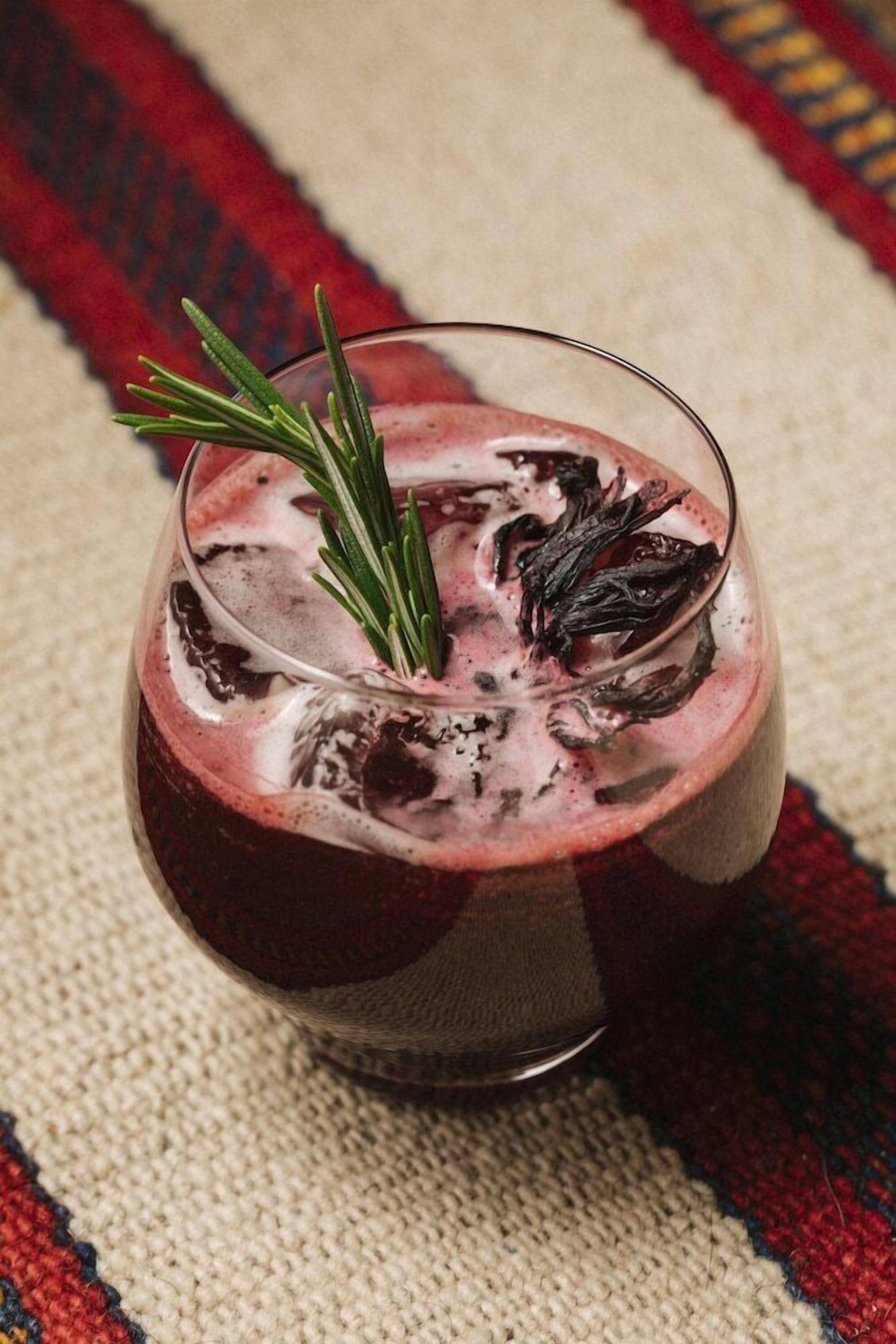 A glass of red beverage with ice and a sprig of rosemary on a textured surface.