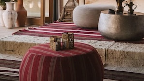 A room with a stone wall, a striped rug, a round ottoman, and a coffee table with decorative items.