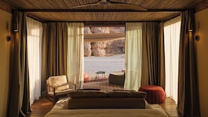 A bedroom with a large bed, a chair, a small table, and a view of a rocky landscape through sheer curtains.
