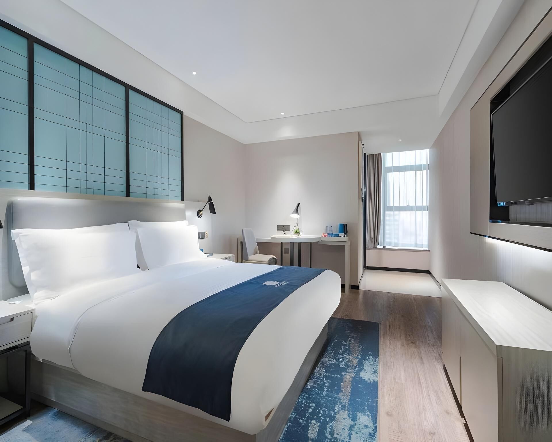 Ein modernes Hotelzimmer mit einem großen Bett, einem Schreibtisch, einem Stuhl, einem Fernseher und einem Fenster mit Vorhängen.