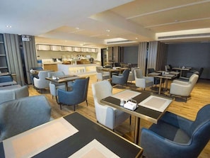 Un hall d’hôtel moderne avec un bar central, des espaces aménagés pour s’asseoir et une cuisine visible au fond.