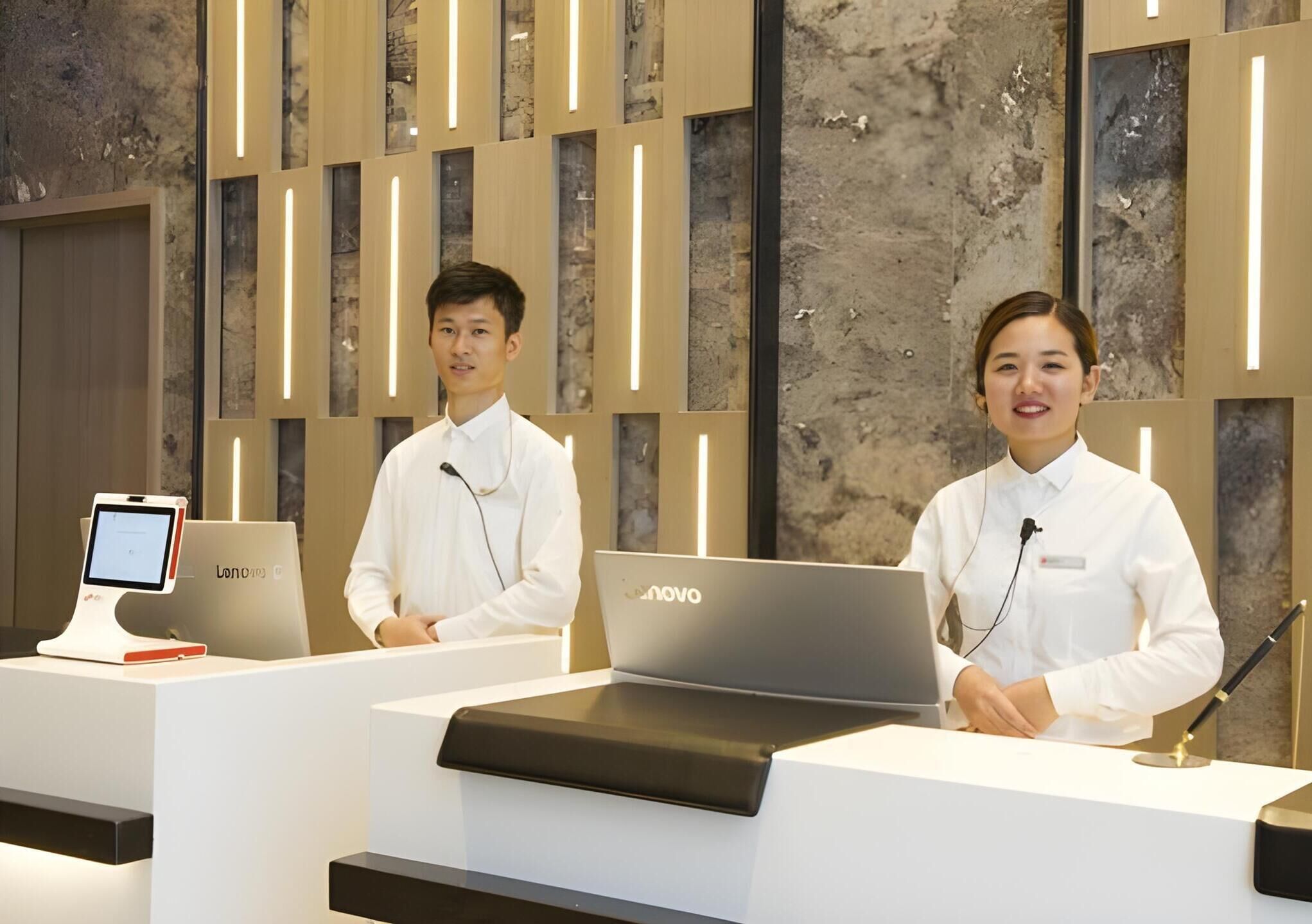 Ein Empfangstresen mit Lenovo-Laptops und einem digitalen Kiosk, vor einer modernen Wand mit vertikalen Holzleisten und einer strukturierten Steinwand.