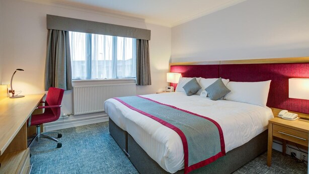 A hotel room with a large bed, a desk with a chair, a window with curtains, and a bedside table with a lamp. - Warwick Conferences - Radcliffe House (Coventry)