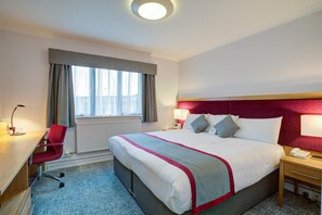 A hotel room with a large bed, a desk with a chair, a window with curtains, and a bedside table with a lamp. - Warwick Conferences - Radcliffe House (Coventry)