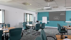 A conference room with teal chairs, a wooden table, a projector, and a screen displaying "Warwick Conferences" and "Hello!". - Warwick Conferences - Radcliffe House (Coventry)