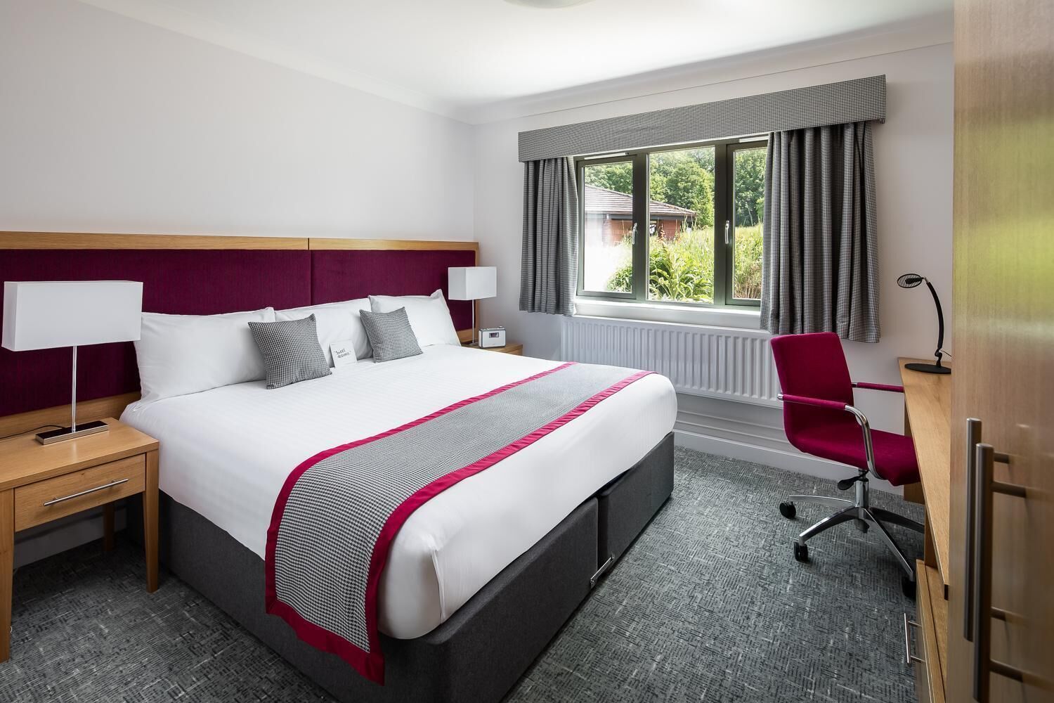 A hotel room with a large bed, a desk, a chair, a nightstand, and a window with curtains.