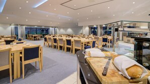 A spacious dining area with multiple tables and chairs, a kitchen area with a bread oven, and large windows providing natural light. - Warwick Conferences - Radcliffe House (Coventry)