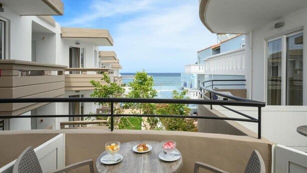 A balcony with a table set for two, overlooking a coastal view with buildings and trees. - Mare Hotel (Apokoronas)