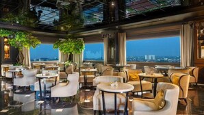A modern lounge with round tables and comfortable seating, large windows with a city view, and a high ceiling with greenery. - Mare Hotel (Apokoronas)