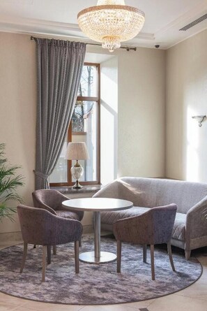 A room with a round table, two chairs, a sofa, a chandelier, and a window with curtains. - Calvary Hotel and Restaurant Vilnius (Vilnius)