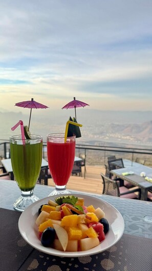 A table with two drinks, a plate of fruit, and a scenic view. - The View (Al Hamra)