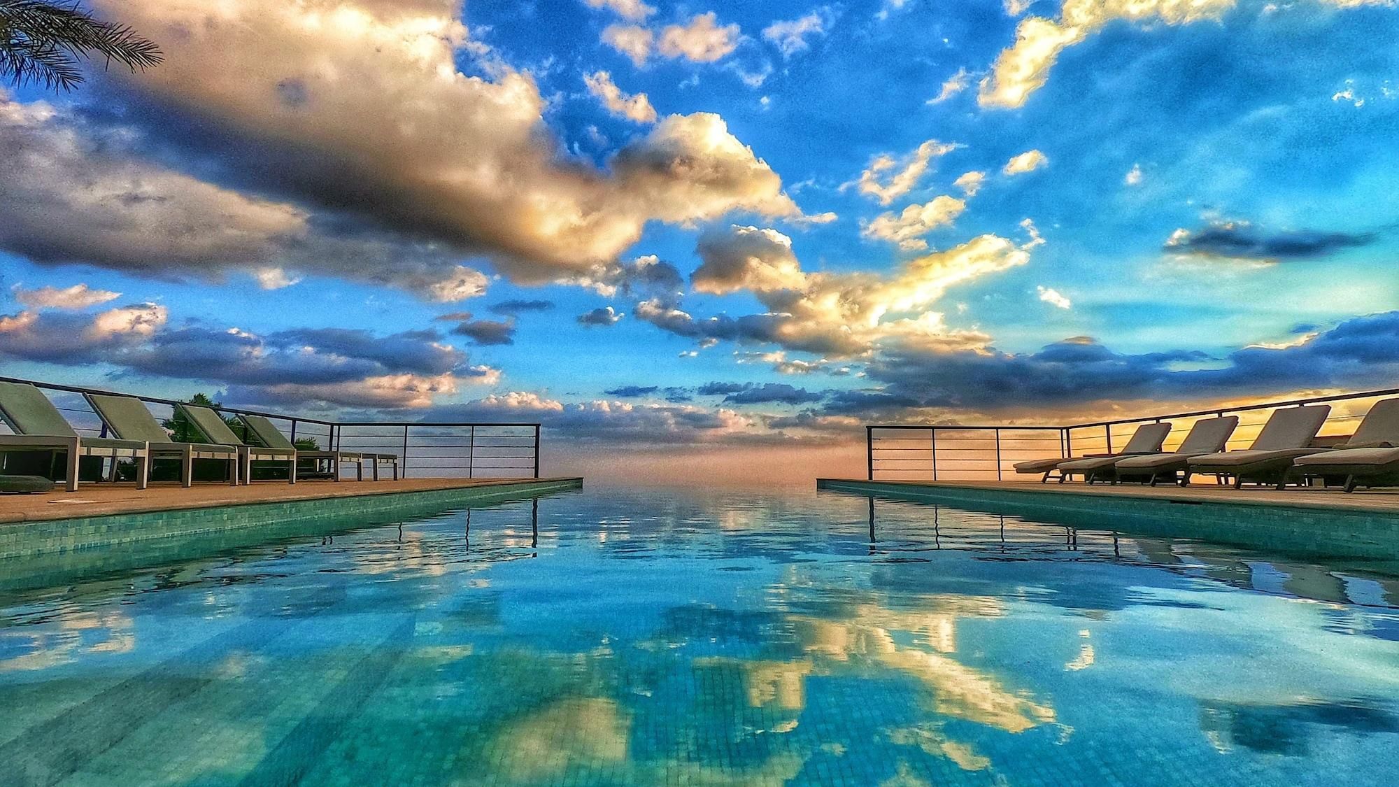 Una piscina infinita con sillones de descanso a ambos lados, bajo un cielo parcialmente nublado.