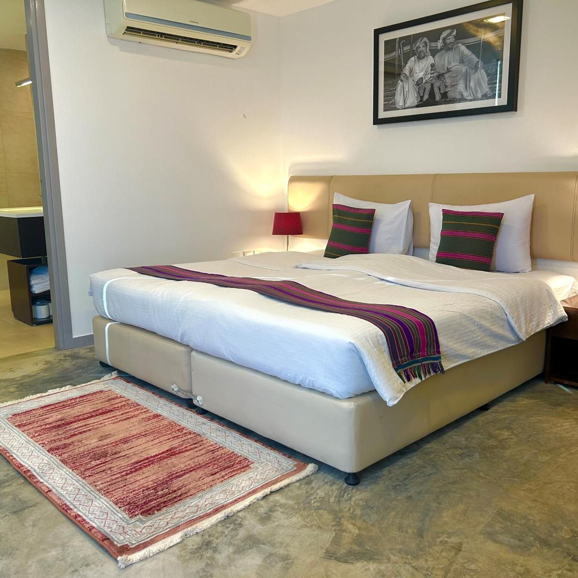 A hotel room with a bed, a nightstand, a lamp, a framed picture on the wall, and a red rug.