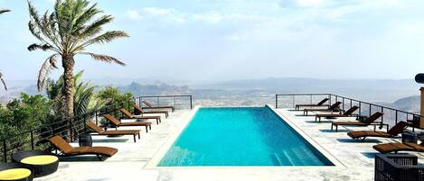 Piscina infinita en la azotea con sillones de descanso y una palmera, con vistas a un paisaje montañoso.