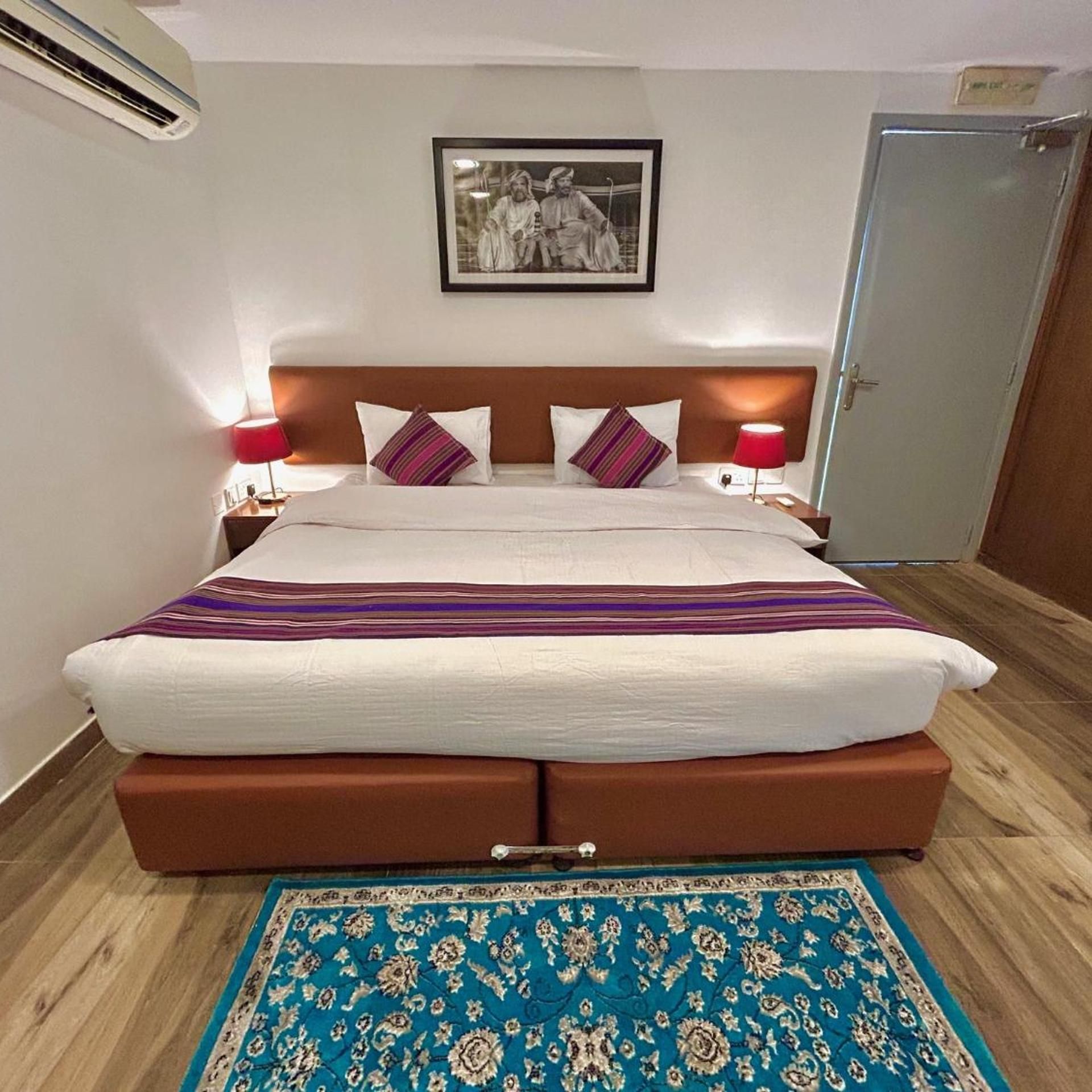 A hotel room with a large bed, two bedside tables, a nightstand, a framed picture on the wall, and a blue patterned rug on the floor.