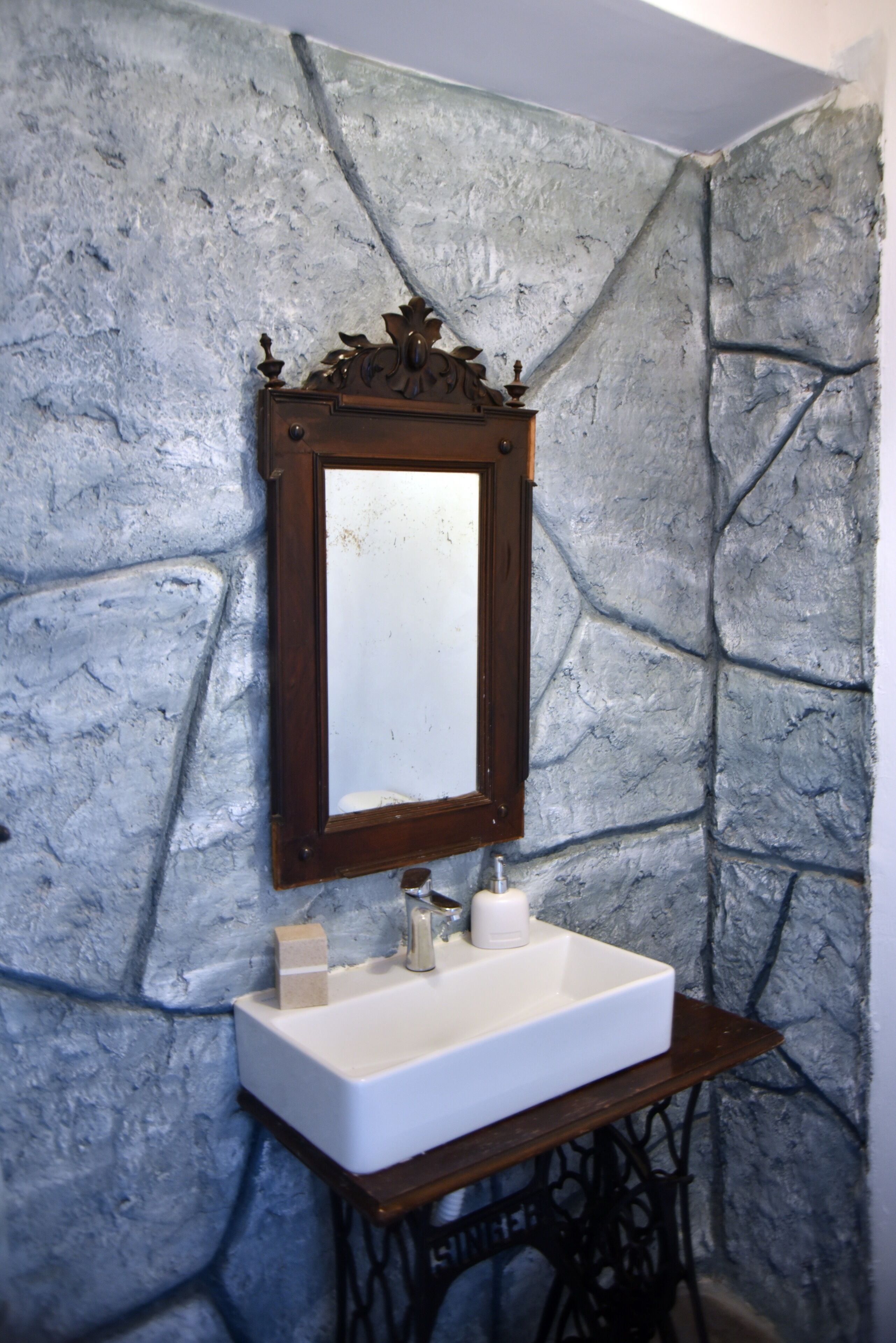 A bathroom with a stone wall, a wooden-framed mirror, and a white sink with soap and a bottle.