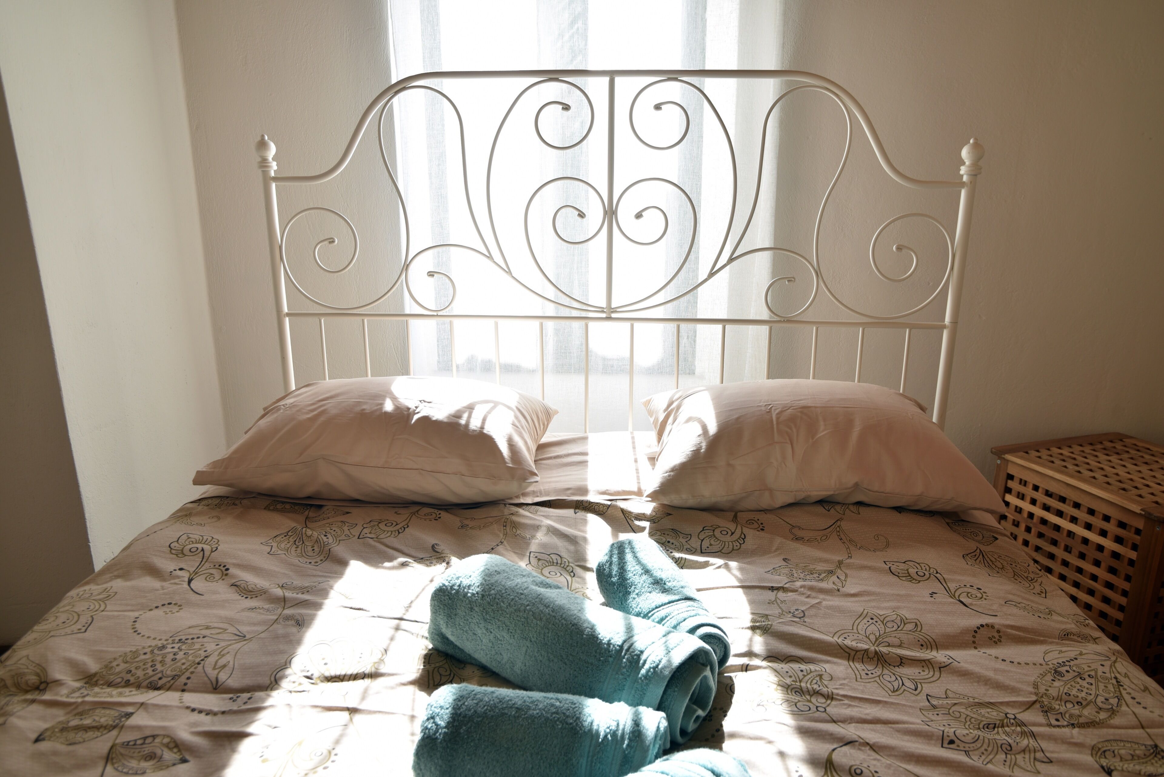 A bed with a metal headboard, two pillows, and a rolled-up towel on a patterned bedspread.