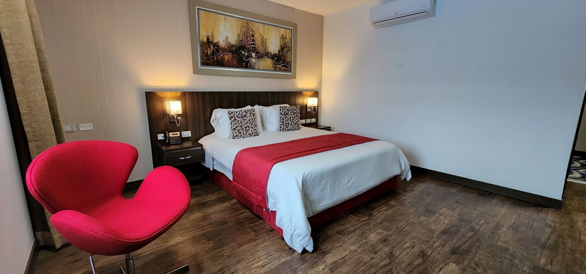 Una camera d'albergo con un letto, una poltrona rossa, un comodino, un quadro e un condizionatore d'aria.