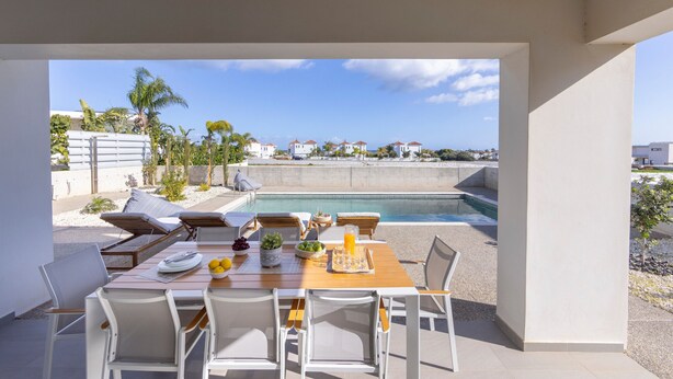 An outdoor dining area with a pool view, white furniture, and a clear sky.