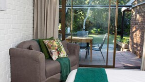 A bedroom with a large bed, a grey sofa with a green throw, and a view of a garden through glass doors. - Tsitsikhaya Lodge (Storms River)