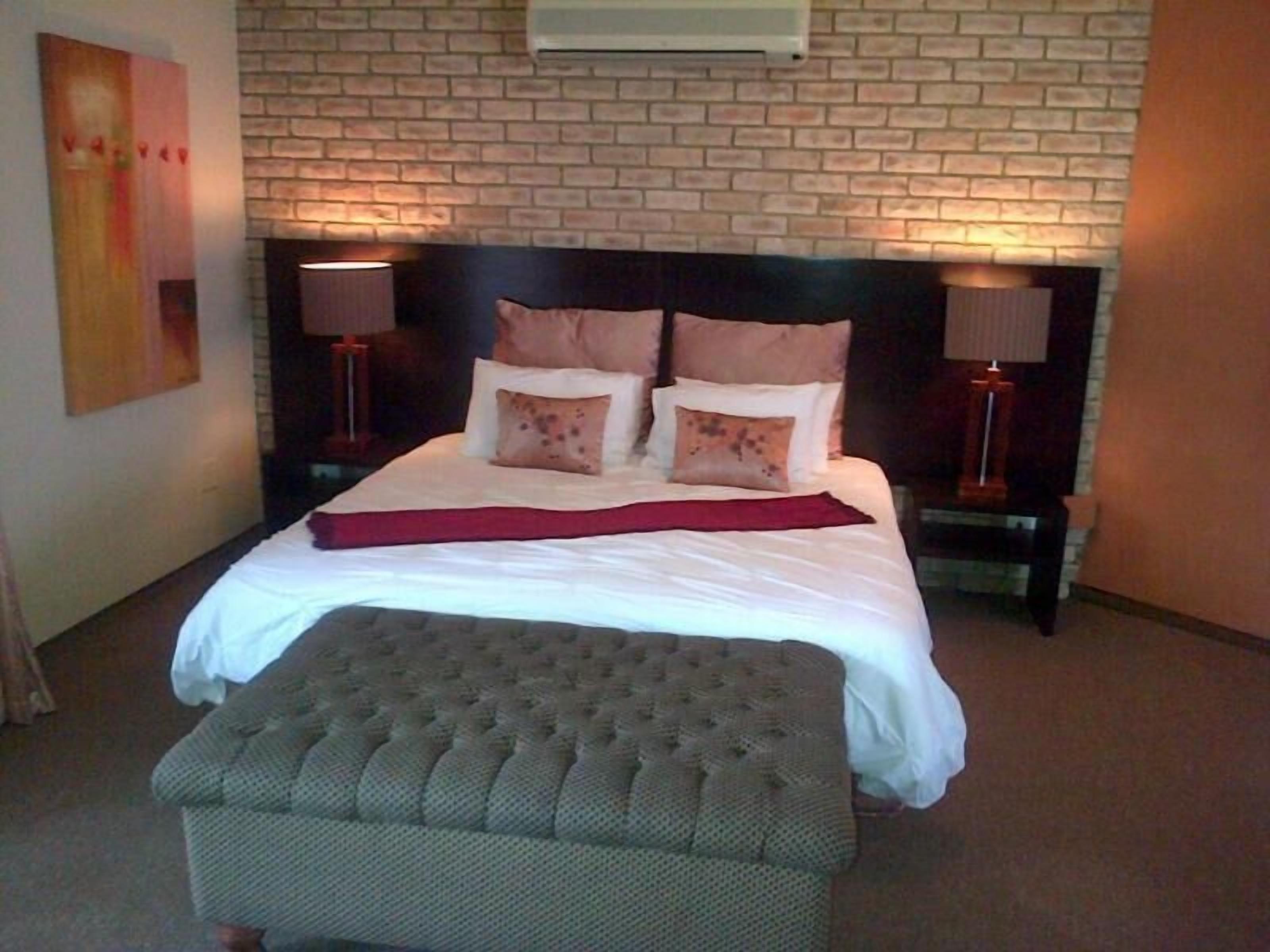 A neatly made bed with a red throw blanket, a headboard with two bedside lamps, and a brick-patterned wall.