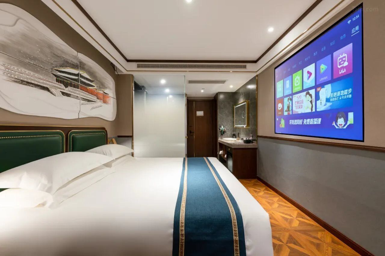 Quarto de hotel com uma cama grande, uma tela digital exibindo diversos aplicativos, um banheiro com espelho e uma parede com pintura de navio.