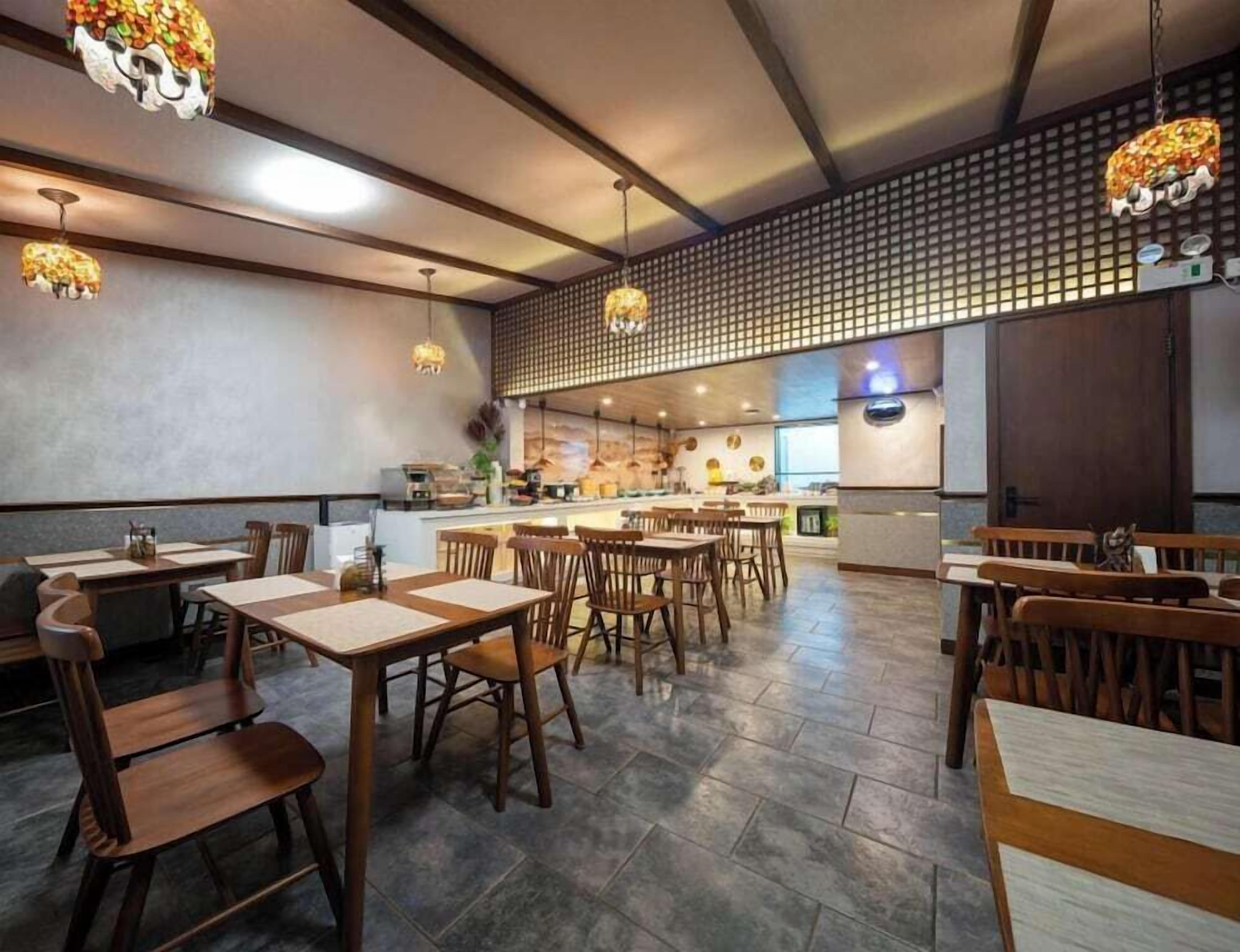 A restaurant interior with wooden tables and chairs, hanging pendant lights, a kitchen area, and a decorative wall.