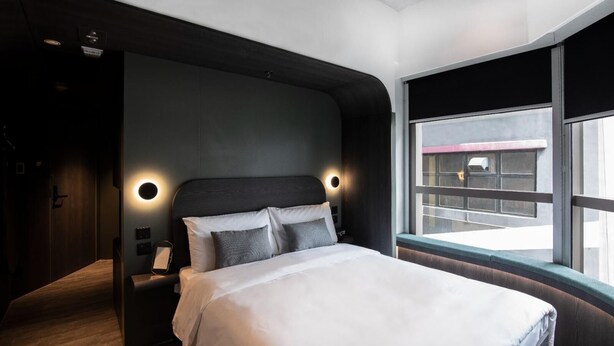 Una habitació d'hotel moderna amb un llit gran, parets fosques i una finestra amb vista a la ciutat.