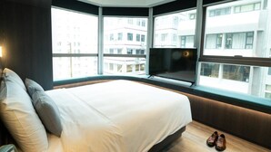 Habitació d'hotel moderna amb un llit gran, una televisió i vista a la ciutat a través de grans finestres.