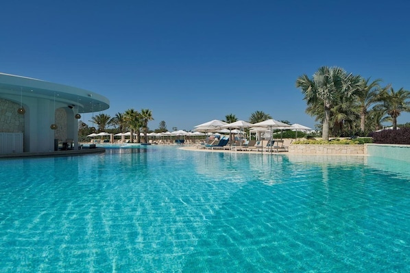 A clear blue pool with lounge chairs and umbrellas, surrounded by palm trees and a modern building.