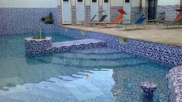 Une piscine avec des marches carrelées menant à l’eau, entourée de chaises longues et d’un bâtiment doté de plusieurs portes.
