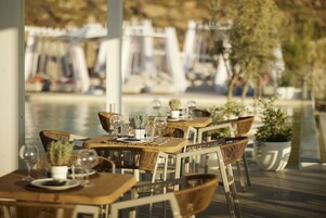 Un área de comedor al aire libre con mesas y sillas de madera, plantas en macetas y vasos de vidrio transparente.