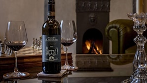 Une bouteille de vin Brunello di Montalcino, deux verres de vin rouge, un jeu d’échecs, une cheminée et une bougie sur une table.