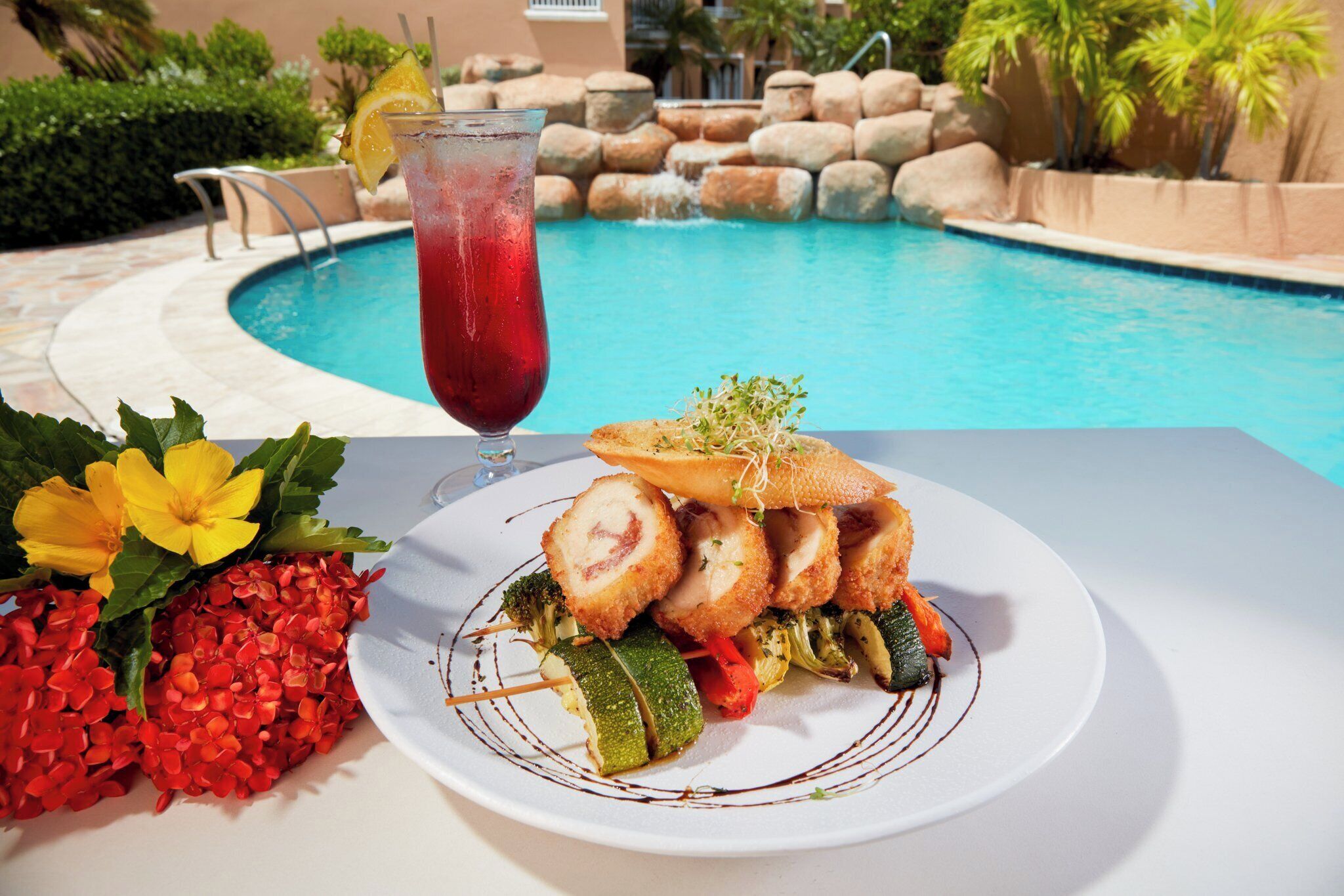 Une table à manger avec une assiette de nourriture, un verre et une piscine en arrière-plan.