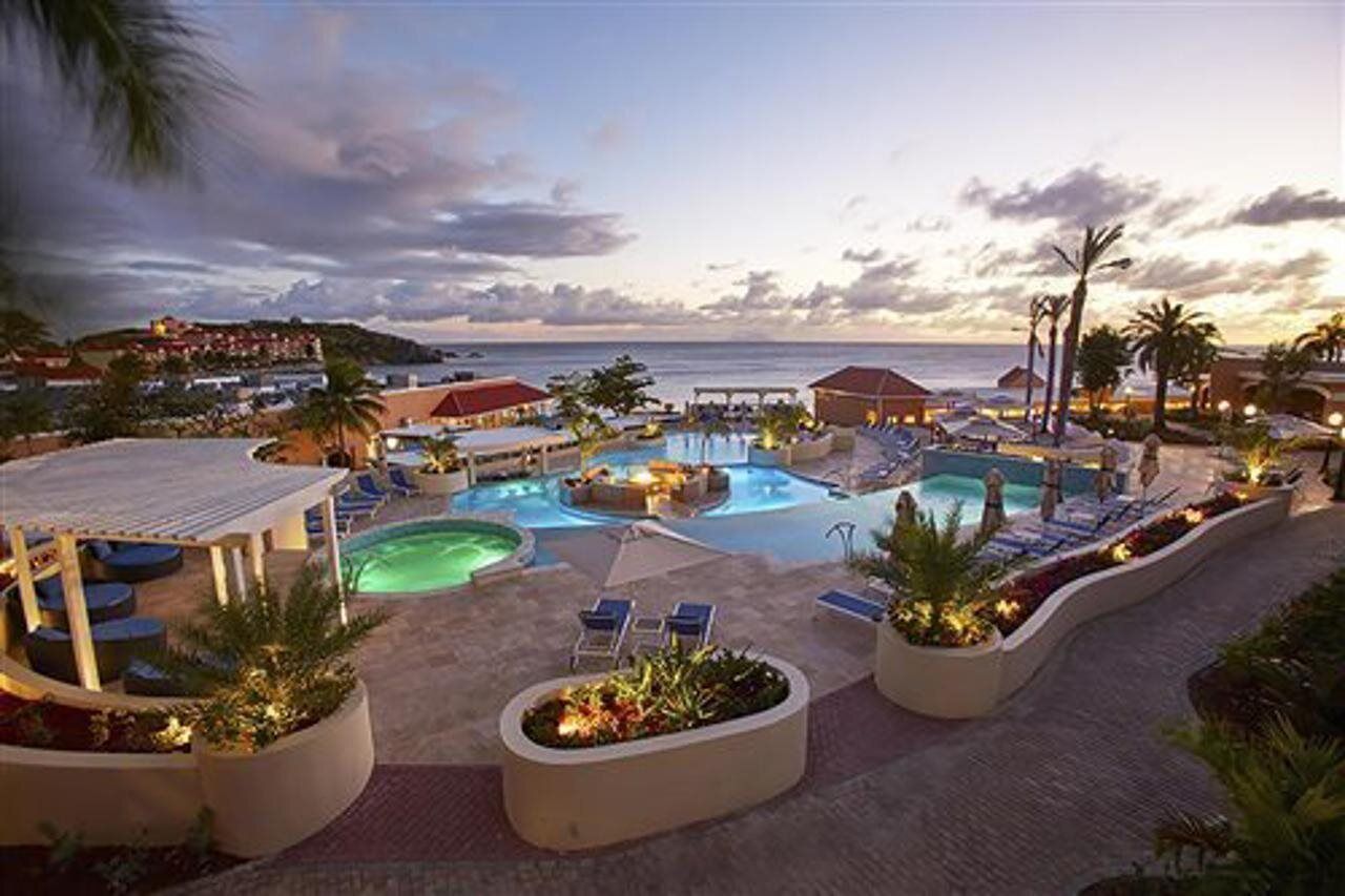 A resort pool area with multiple pools, lounge chairs, and a view of the ocean at dusk.