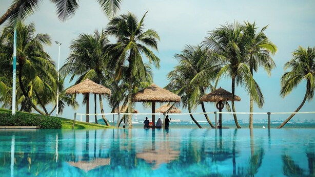 A swimming pool with palm trees and thatched umbrellas, surrounded by a clear view of the ocean. - Forest City Eco Stay (Gelang Patah)
