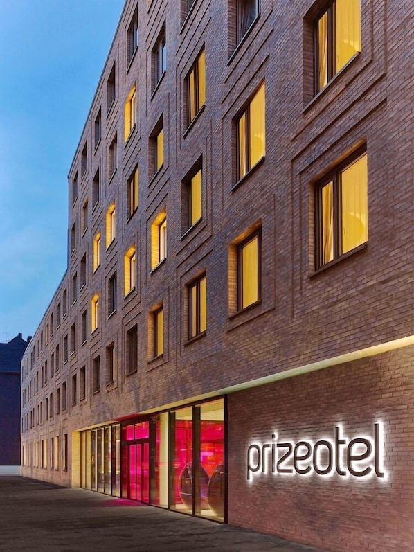 Fachada moderna de un hotel con un llamativo letrero de "prizeotel", con revestimiento de ladrillo y múltiples ventanas con vidrios tintados de amarillo.