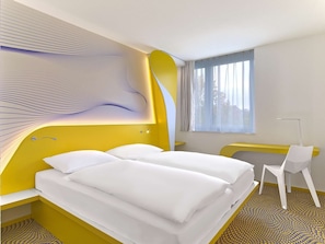 Ein modernes Hotelzimmer mit gelber Akzentwand, einem großen Bett, einem Schreibtisch und einem gemusterten Boden.