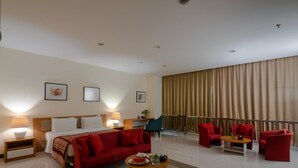 Una habitación de hotel moderna con una cama, sofá rojo, una mesa de comedor con un plato de comida y un ventanal con cortinas.