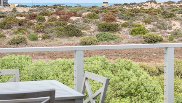 Une terrasse en bois avec une table et des chaises, donnant sur une plage à la végétation clairsemée et sous un ciel dégagé.