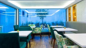 Un restaurante moderno con sillas tapizadas en verde y mesas blancas, ventanas amplias que ofrecen vistas al exterior y un árbol decorativo en el centro.